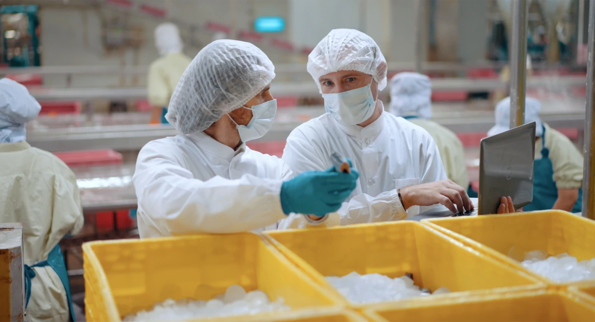 Food processing workers in protective clothing and face masks discussing quality checks beside iced ingredient storage bins on a commercial production floor.